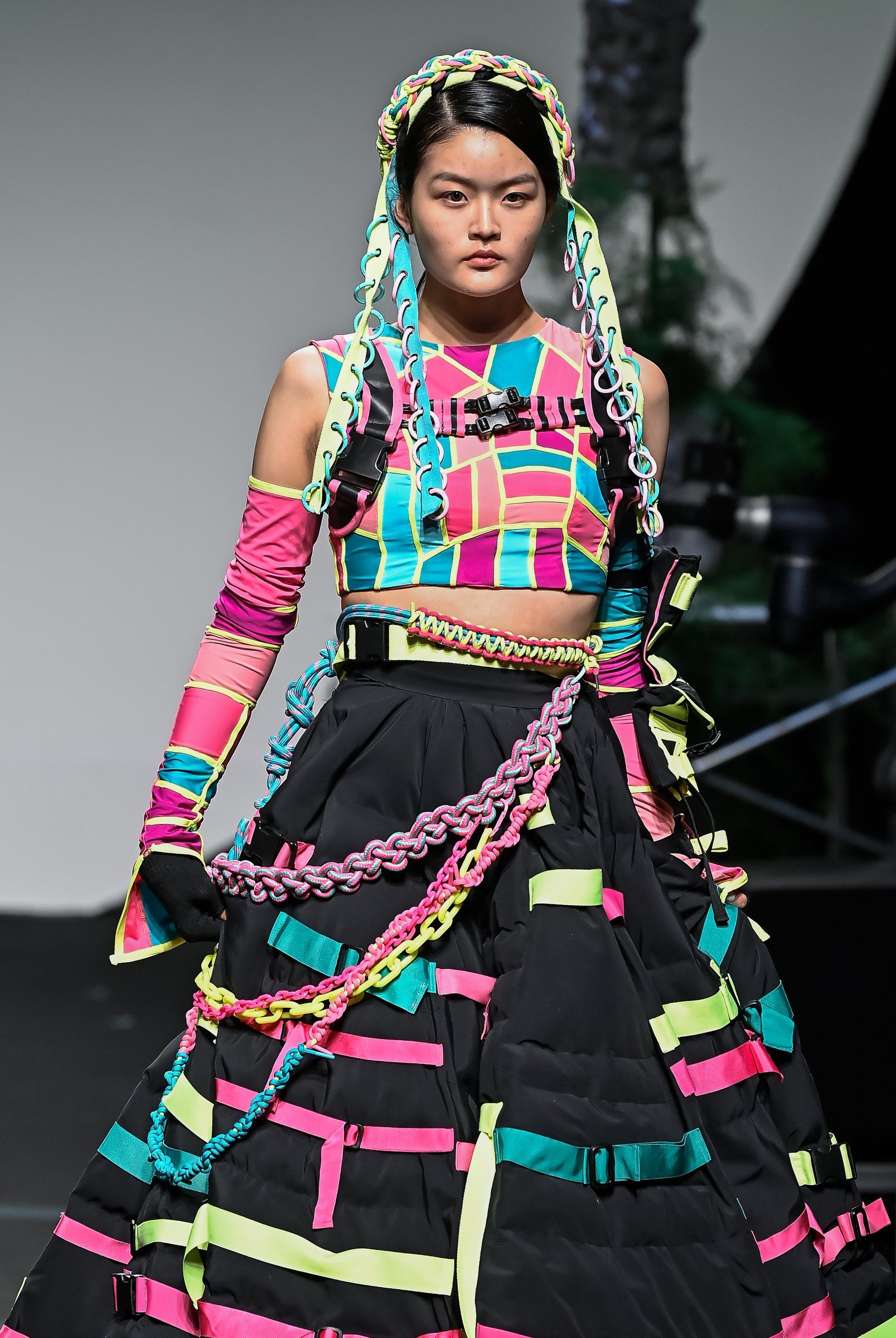 Model on the runway in a patchwork lycra crop top with a hooded braid and a black quilted ball skirt striped with lime, pink and cyan tape